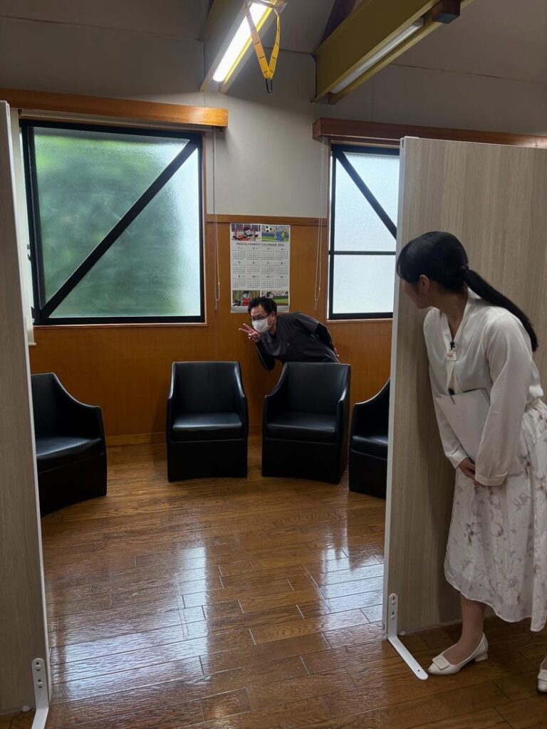 診察室の椅子の奥から、顔をのぞかせてピースサインをする恵川先生