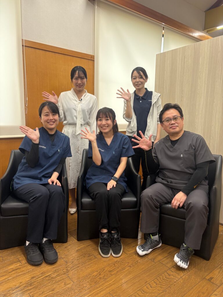 室内で、椅子に座った3名と、その後ろに立つ2名のスタッフが、手を振りながら並んでいる集合写真