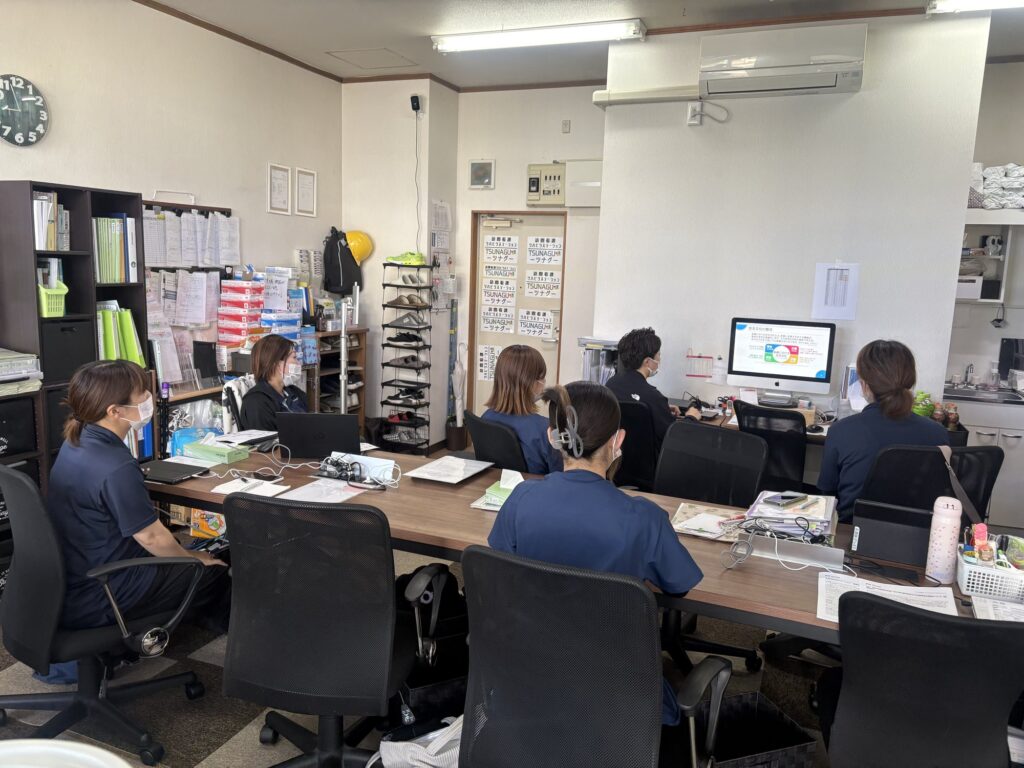 事務所内で、スタッフが長机を囲んで座り、モニターに映る資料を見ながら研修や情報共有をしている様子